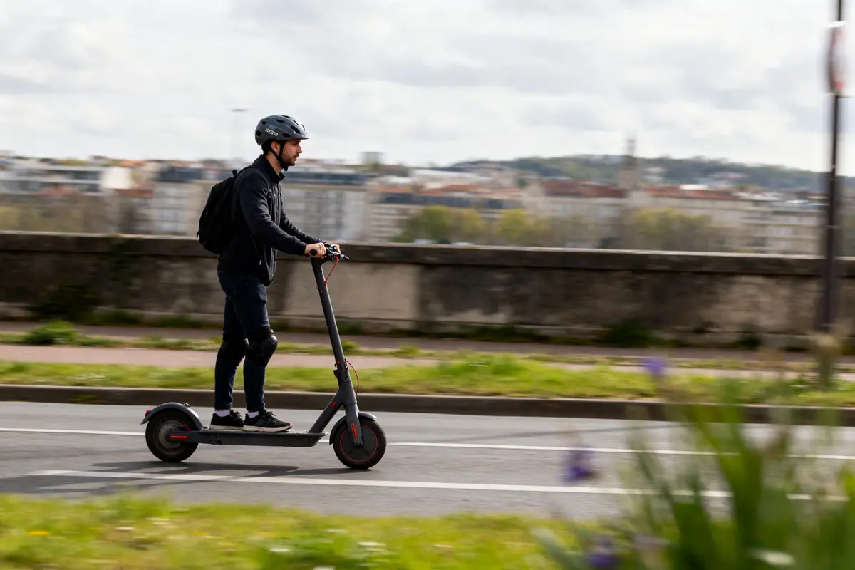 Un homme roule en trottinette électrique, portant un casque et un sac à dos en ville.
