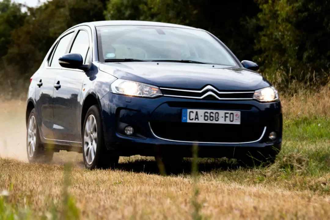 Voiture Citroën bleue roulant sur chemin herbeux, arbres en arrière-plan, immatriculation visible.