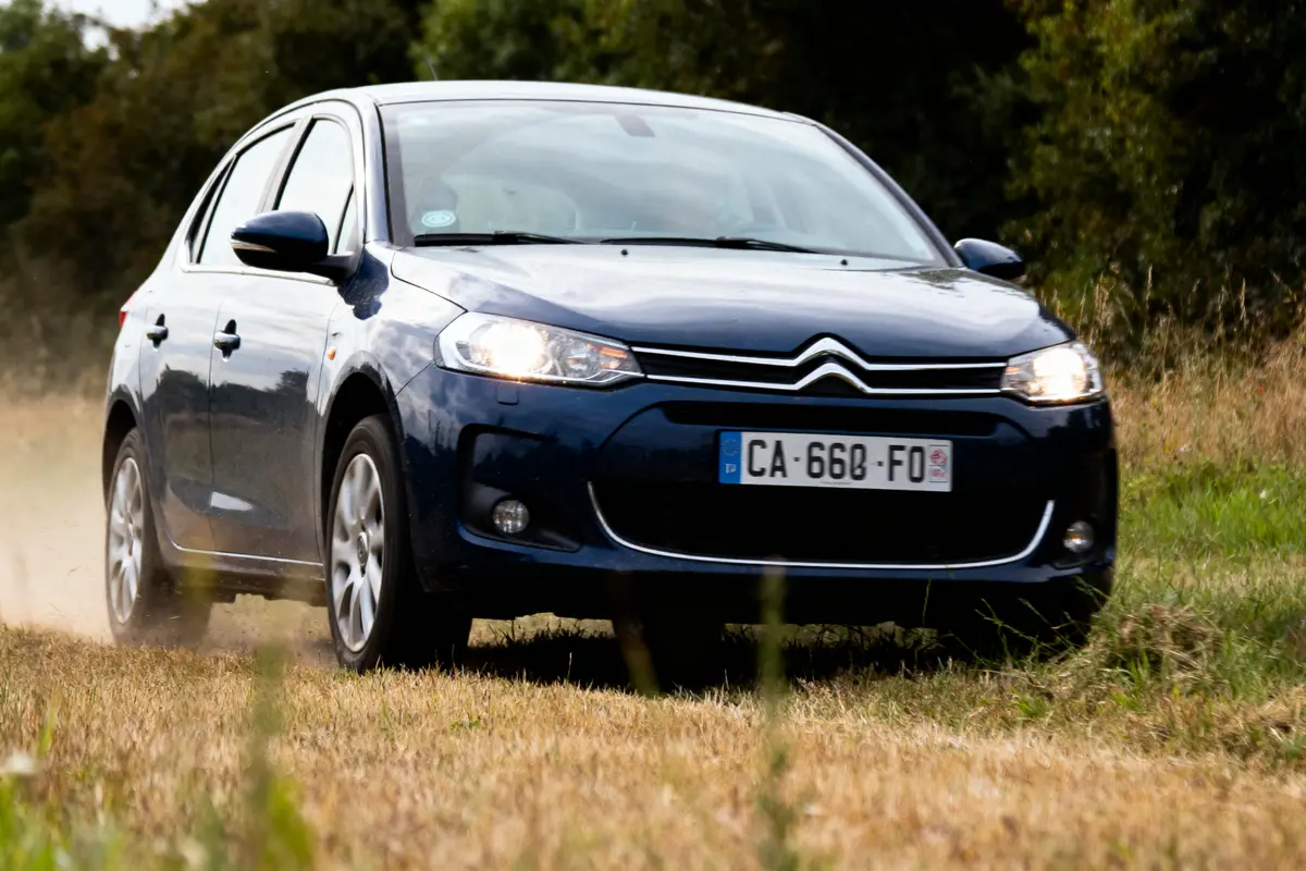 Voiture Citroën bleue roulant sur chemin herbeux, arbres en arrière-plan, immatriculation visible.