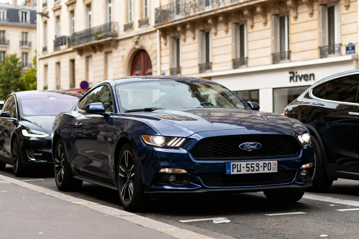 Voiture Ford bleue garée en ville, entre deux autres véhicules, devant un bâtiment urbain.