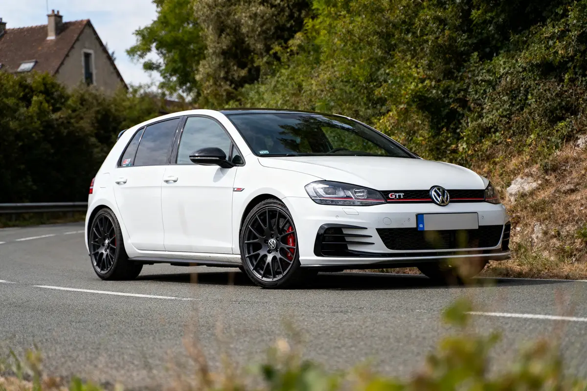 Voiture blanche Volkswagen GTI sur route sinueuse bordée de végétation et maison en arrière-plan.